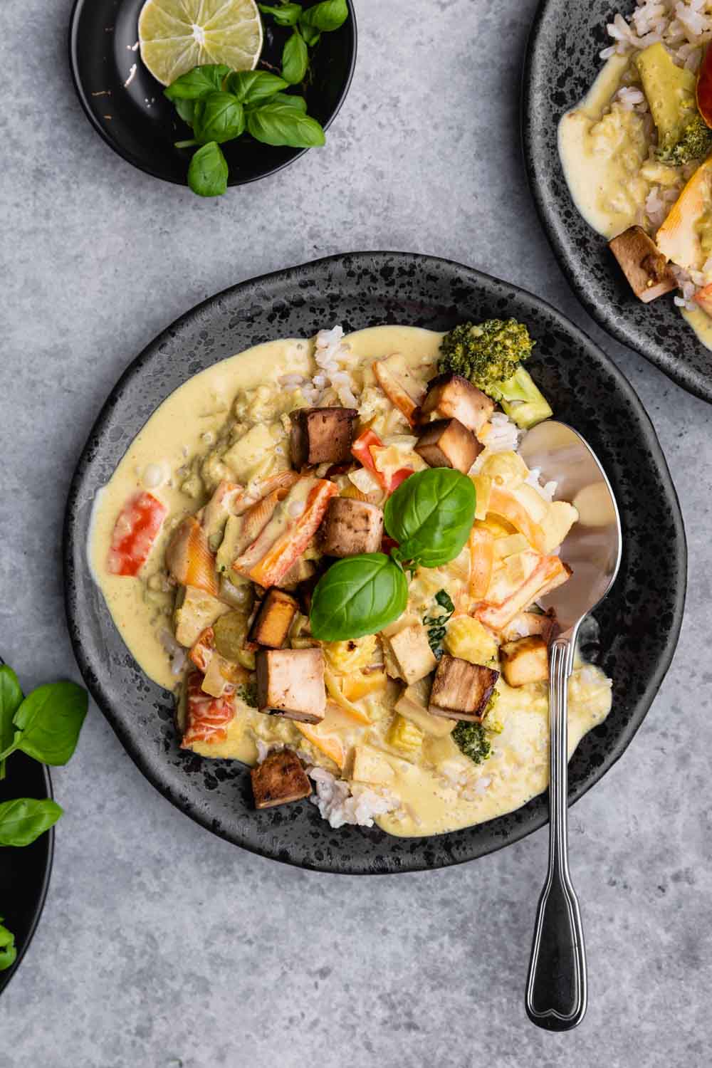 Green curry tofu in a black bowl with tofu and a basil leaf on top and a silver spoon on a grey backdrop