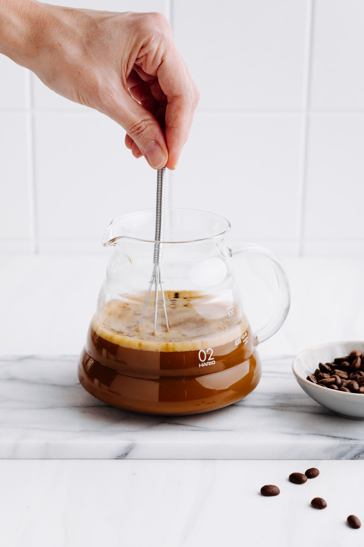 A hand with a whisk in a glass pot of coffee with milk with coffee beans next to it on a white backdrop