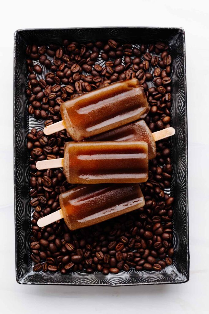 Four brown coffee popsicles on a backdrop of coffee beans in a silver tray on a white backdrop