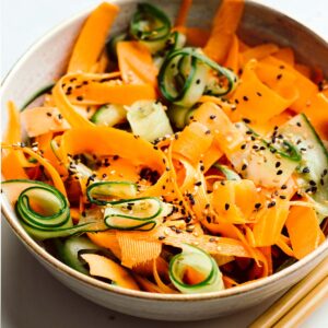 Cucumber and carrot salad sprinkled with white and black sesame seeds in a light brown bowl on a white plate with two wooden chopsticks