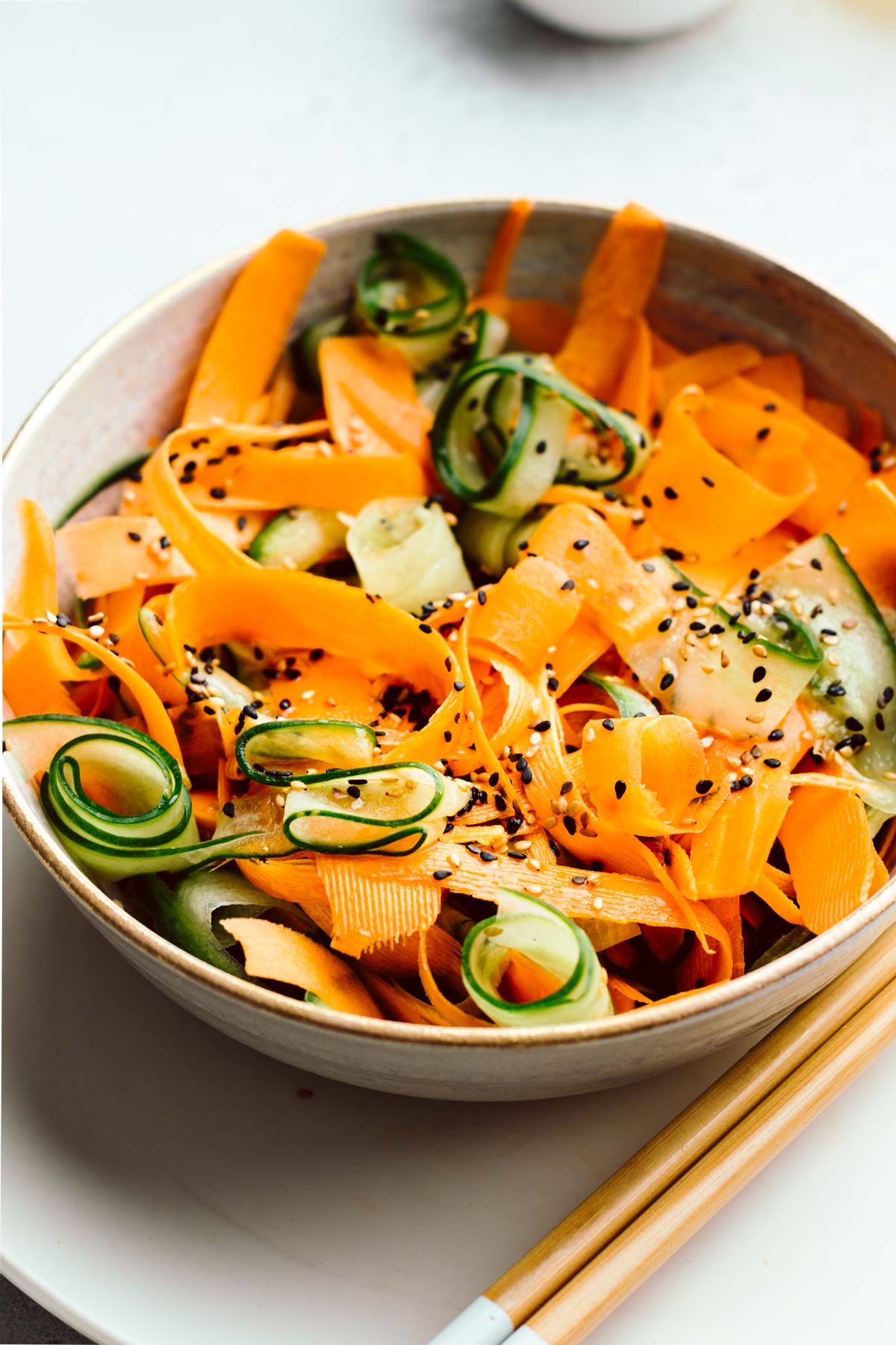 Cucumber and carrot salad sprinkled with white and black sesame seeds in a light brown bowl on a white plate with two wooden chopsticks