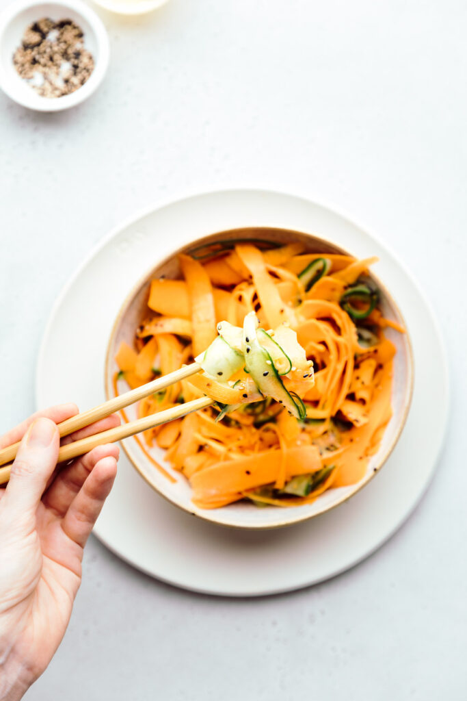 Cucumber and carrot ribbons with the dressing in a light brown bowl on a light grey backdrop and a hand showing some salad ribbons with wooden chopsticks above the bowl