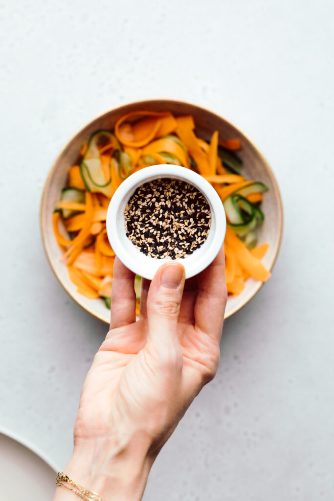 Cucumber and carrot ribbons with the dressing in a light brown bowl on a light grey backdrop and a hand showing a small bowl with sesame seeds above the bowl