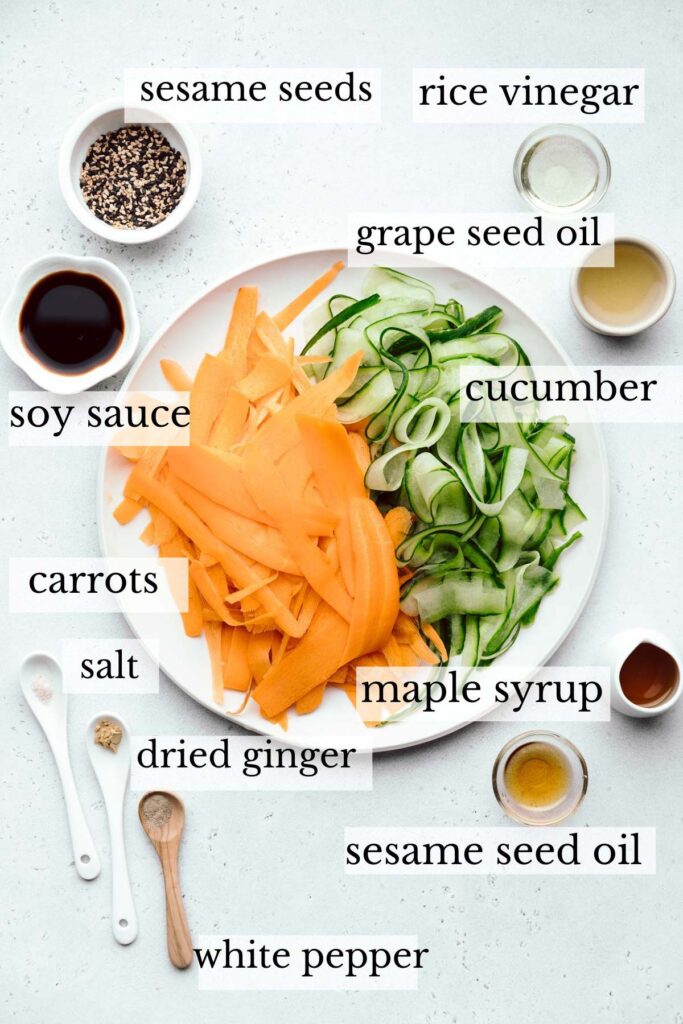 Ingredients for cucumber and carrot salad on a light grey backdrop in separate bowls and spoons and a large plate in the middle