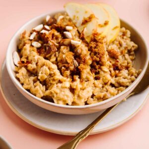 Brown Sugar Cinnamon Oatmeal topped with sliced apples, chopped almonds and brown sugar in a bowl on a plate with a gold colored spoon next to it and a glass of milk in the background.