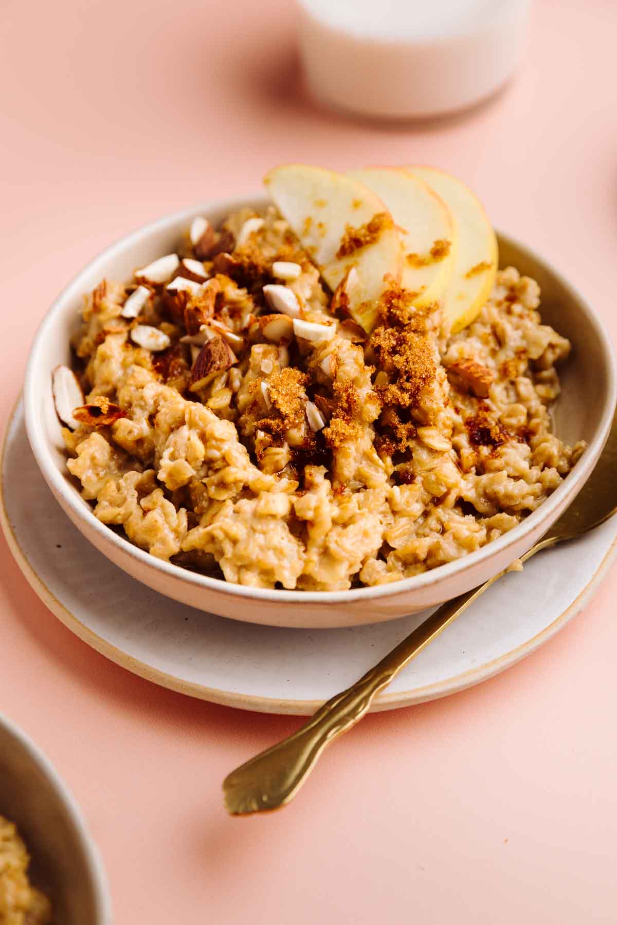 Brown Sugar Cinnamon Oatmeal topped with sliced apples, chopped almonds and brown sugar in a bowl on a plate with a gold colored spoon next to it and a glass of milk in the background.