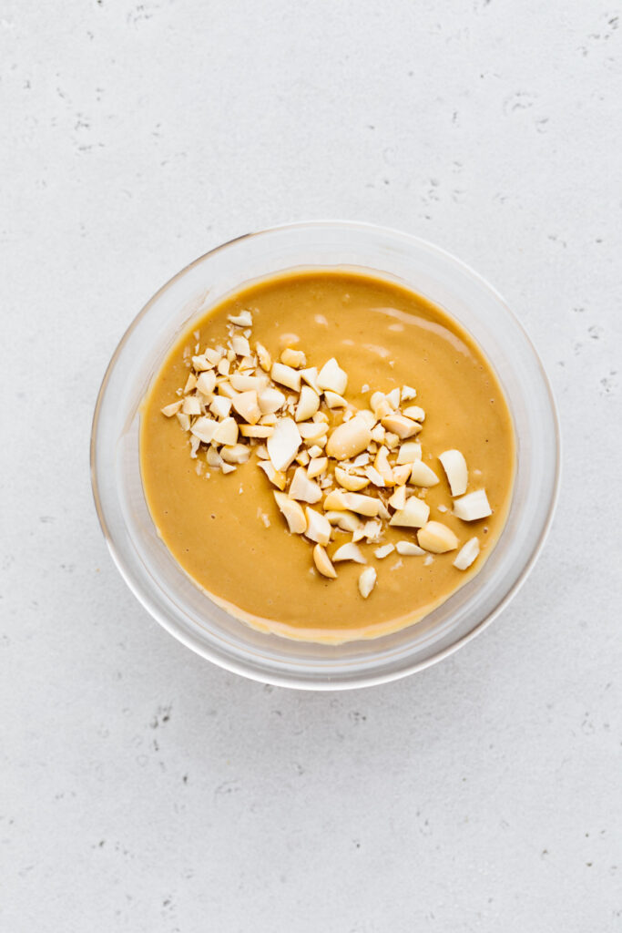 A small glass bowl on a grey backdrop with creamy peanut sauce and garnished with chopped peanuts