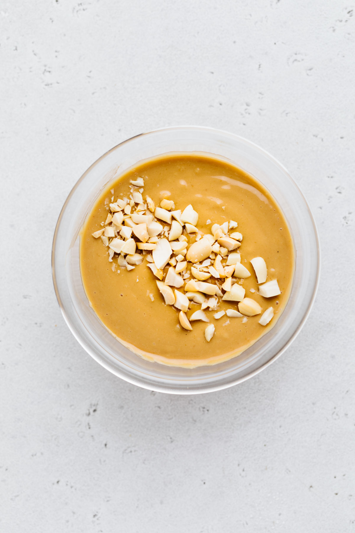 A small glass bowl on a grey backdrop with creamy peanut sauce and garnished with chopped peanuts