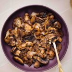A purple plate on a tiled backdrop with caramelized onions and mushrooms with a golden spoon in it and slices of bread showing in the top corner.