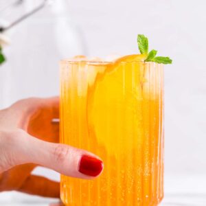 A hand holding a glass with yellow Crodino Spritz with ice cubes garnished with mint and an orange slice on a white marble backdrop.