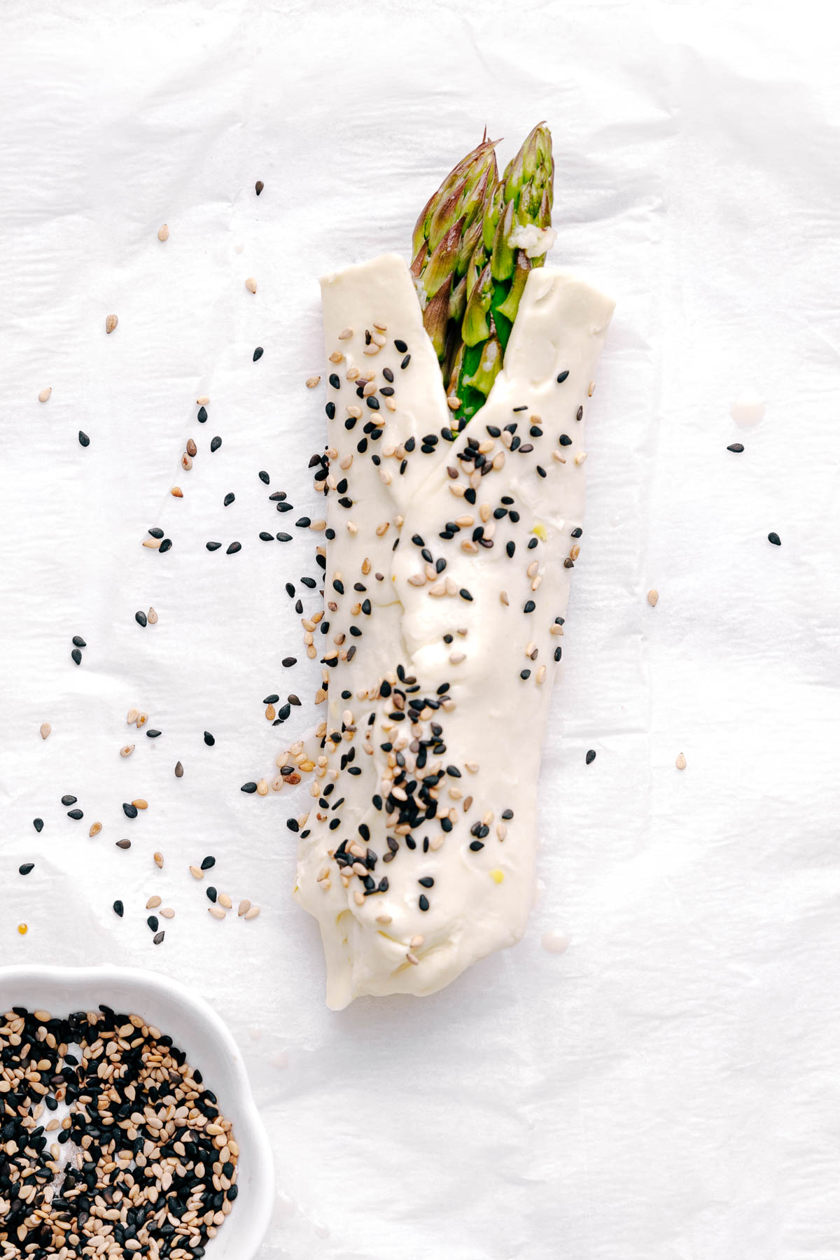 A bundle of Asparagus with Puff Pastry on white parchment paper sprinkled with white and black sesame seeds next to a small bowl with sesame seeds