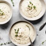 three bowls of creamy cauliflower soup on a grey backdrop in a light bowl decorated with thyme