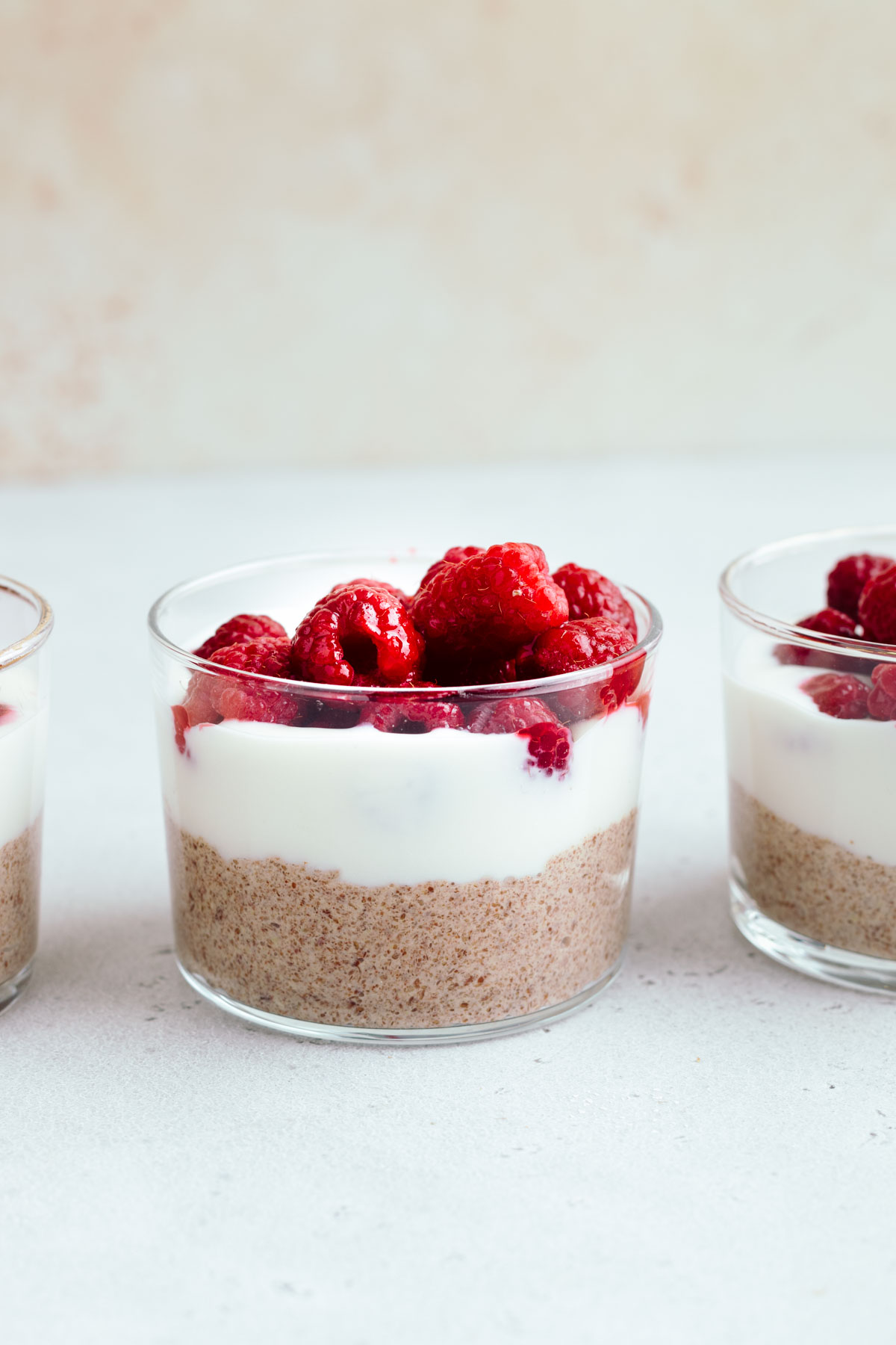 A small glass layered with a light brown pudding, a small layer of white yogurt and fresh raspberries on a light grey backdrop in front of light brown wall next to two small glasses with pudding.