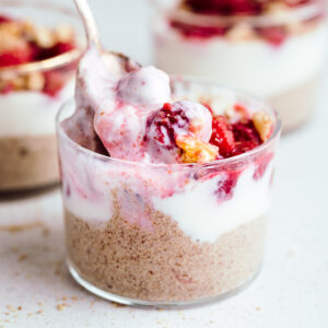 A small glass on a light backdrop with more puddings showing in the back layered with brown pudding, then white yogurt and then red raspberries with chopped walnuts and a spoon scooping up the yogurt with raspberries.