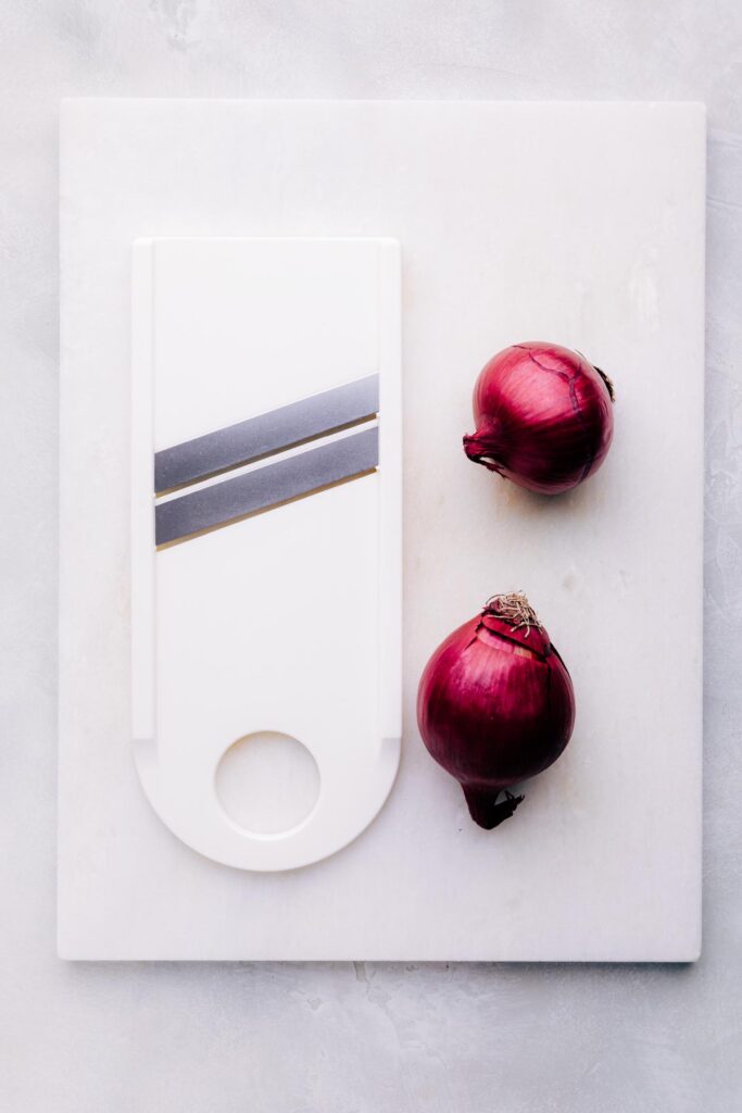 Two red onions and a white mandoline on a white marble cutting board.
