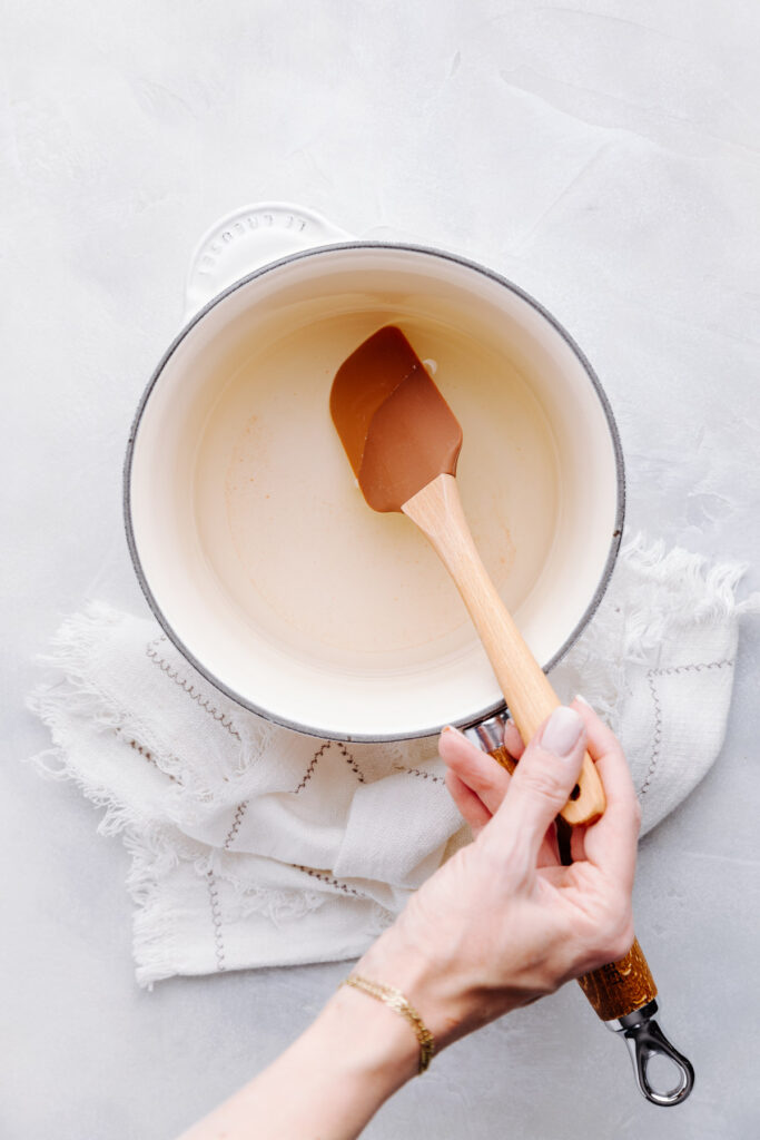 Ingredients to make the brine for pickled red onions in a vanilla colored saucepan on a white napkin with a brown wooden spatula in the saucepan.