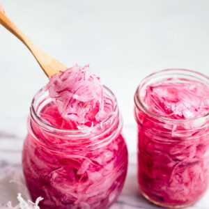 Two glass jars with pickled red onions faced open on a marble cutting board with a wooden fork taking out some pickled red onions