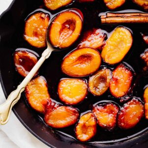Orange and red color plum halves in a syrupy liquid with the inside facing up in a black skillet with a golden spoon with one plum halve in it.