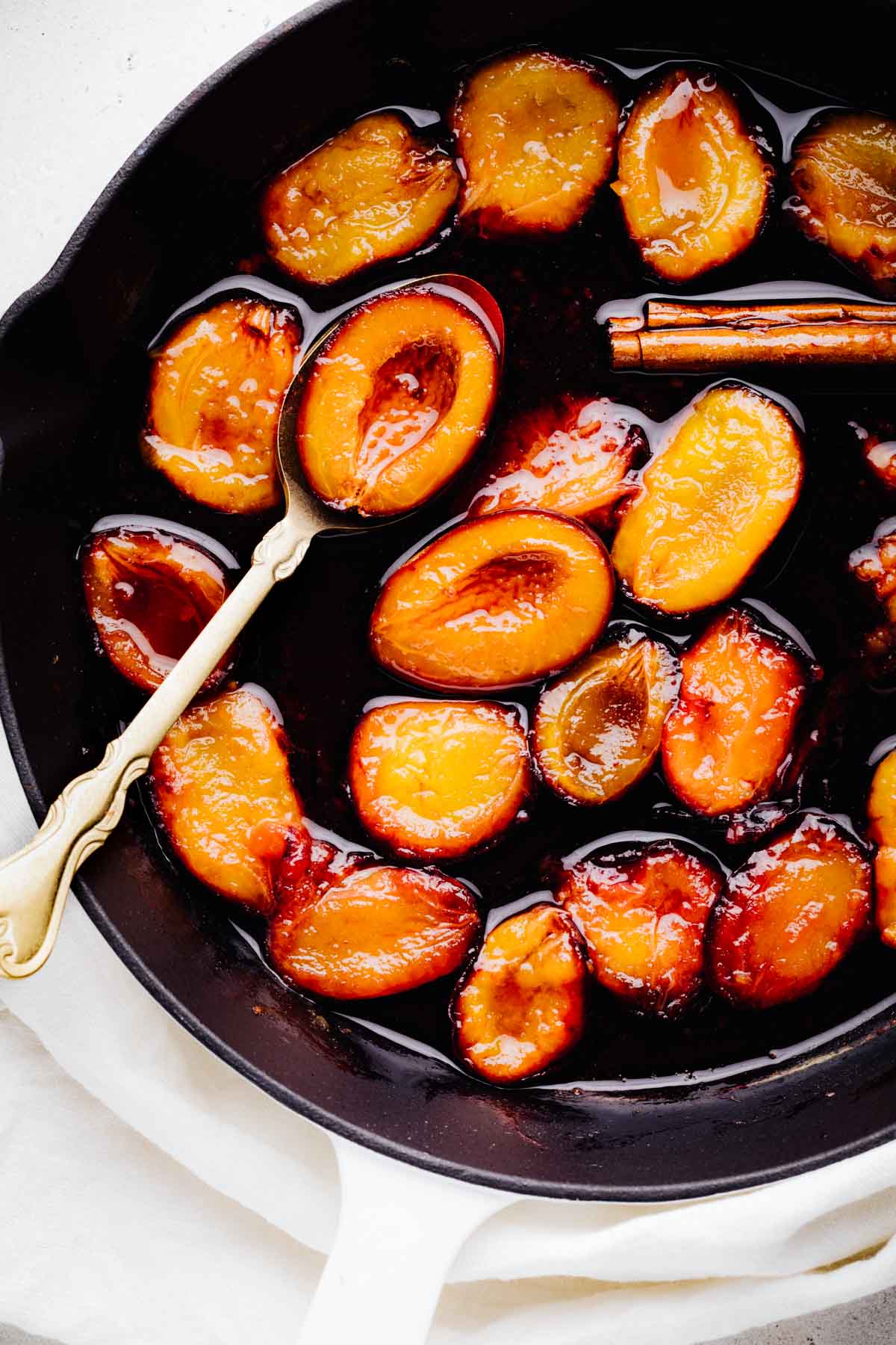 Orange and red color plum halves in a syrupy liquid with the inside facing up in a black skillet with a golden spoon with one plum halve in it.