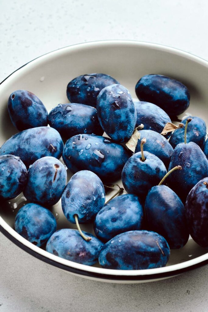 A light grey backdrop with a light colored shallow bowl with a blue edges and bright blue plums with a few water drops on top.