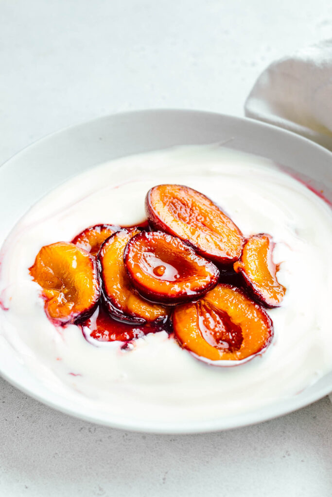 A light grey bowl on a white backdrop with white yogurt in it topped with red and dark orange glossy stewed plum halves and a part of a white napkin in the top right corner.
