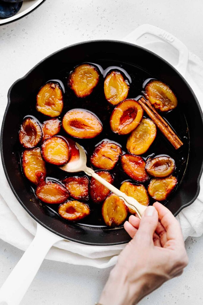 A white skillet with black interior with red and dark yellow stewed plum halves and a cinnamon stick in a glossy liquid and a hand holding a golden spoon in it on a white napkin on a light backdrop.