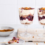 Two tall glasses with vegan parfait on a white wooden board with a wooden spoon next to it and a small bowl with crumbled cookies.