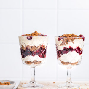 Two tall glasses with vegan parfait on a white wooden board with a wooden spoon next to it with crumbled cookies.
