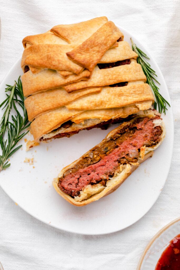 baked vegan wellington on a white plate with rosemary and one slice cut