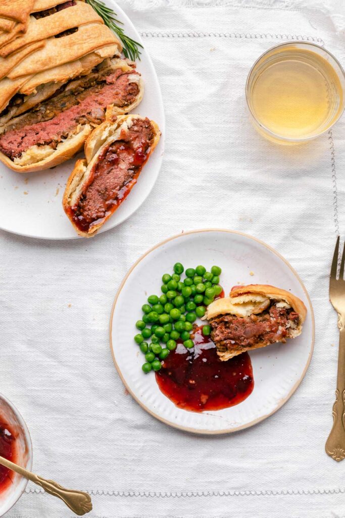 cut baked vegan wellington on a white plate with green peas and cranberry sauce on a white tablecloth