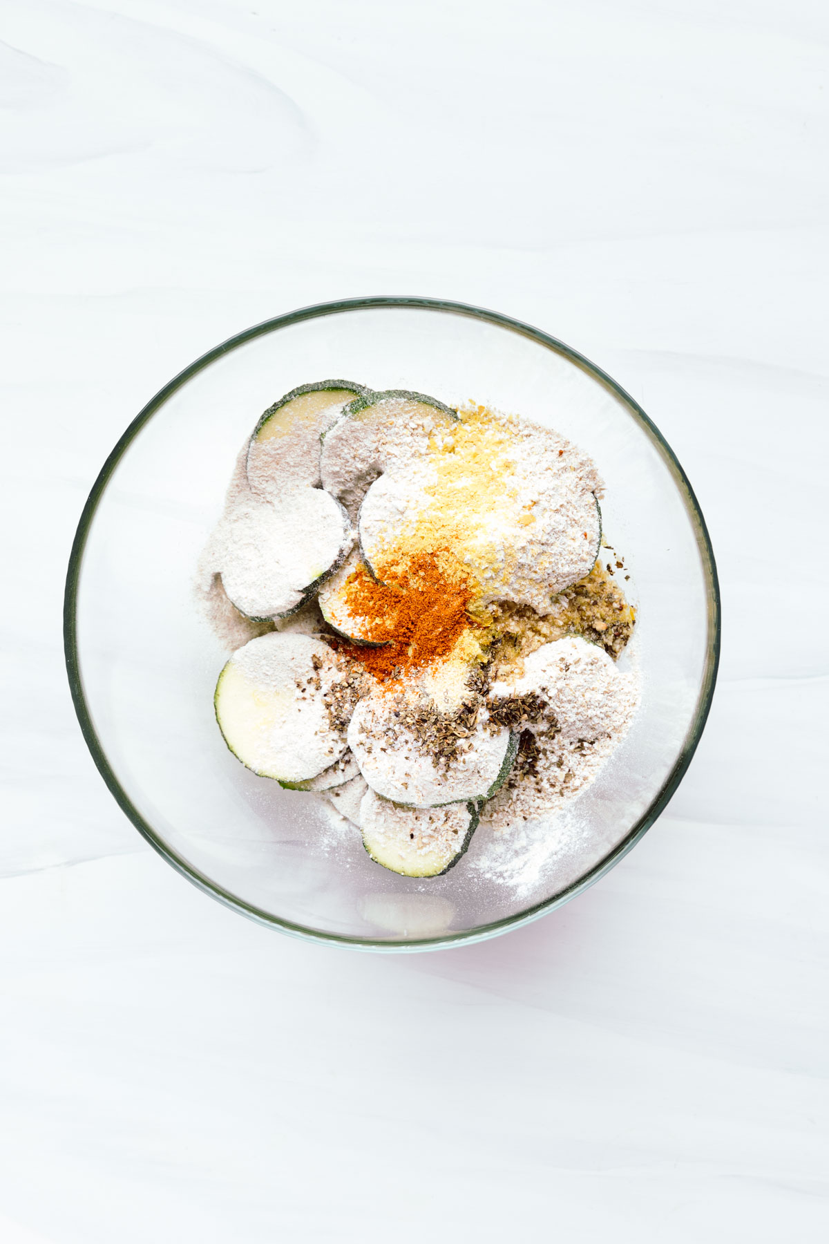Zucchini slices in a glass bowl with flour and paprika powder on top on a white backdrop