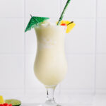 Piña Colada Mocktail in a tall hurricane glass decorated with a green paper umbrella, a pineapple chunk, a cherry and a green paper straw on a marble cutting board on a white backdrop