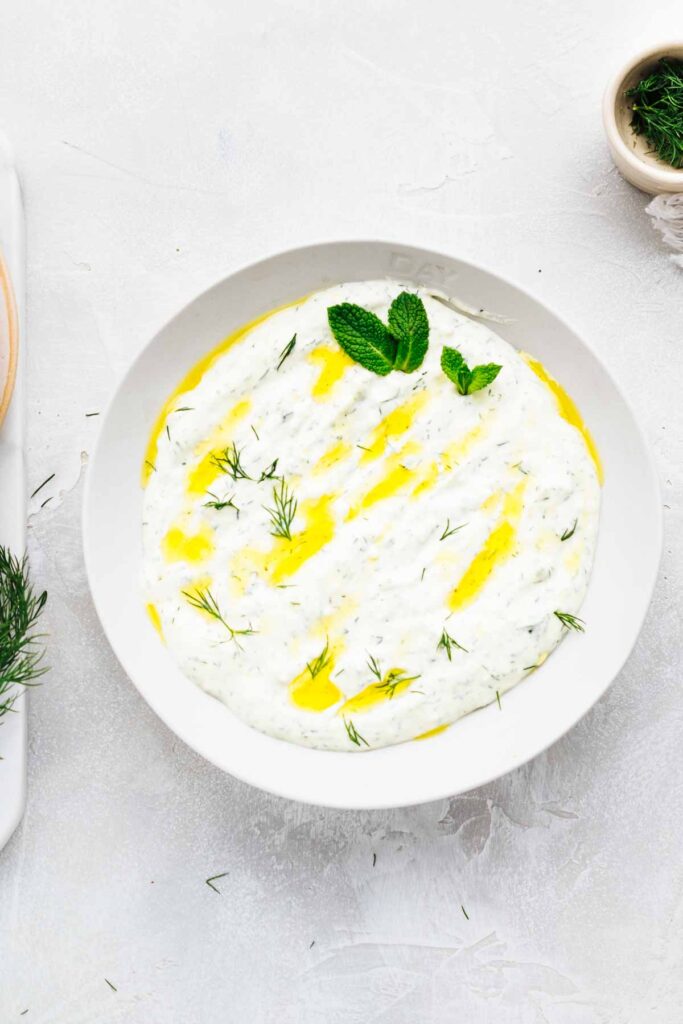 A bowl on a light grey backdrop with a thick dip in it garnished with olive oil, dill and mint leaves and a small bowl with fresh dill in the top right corner.