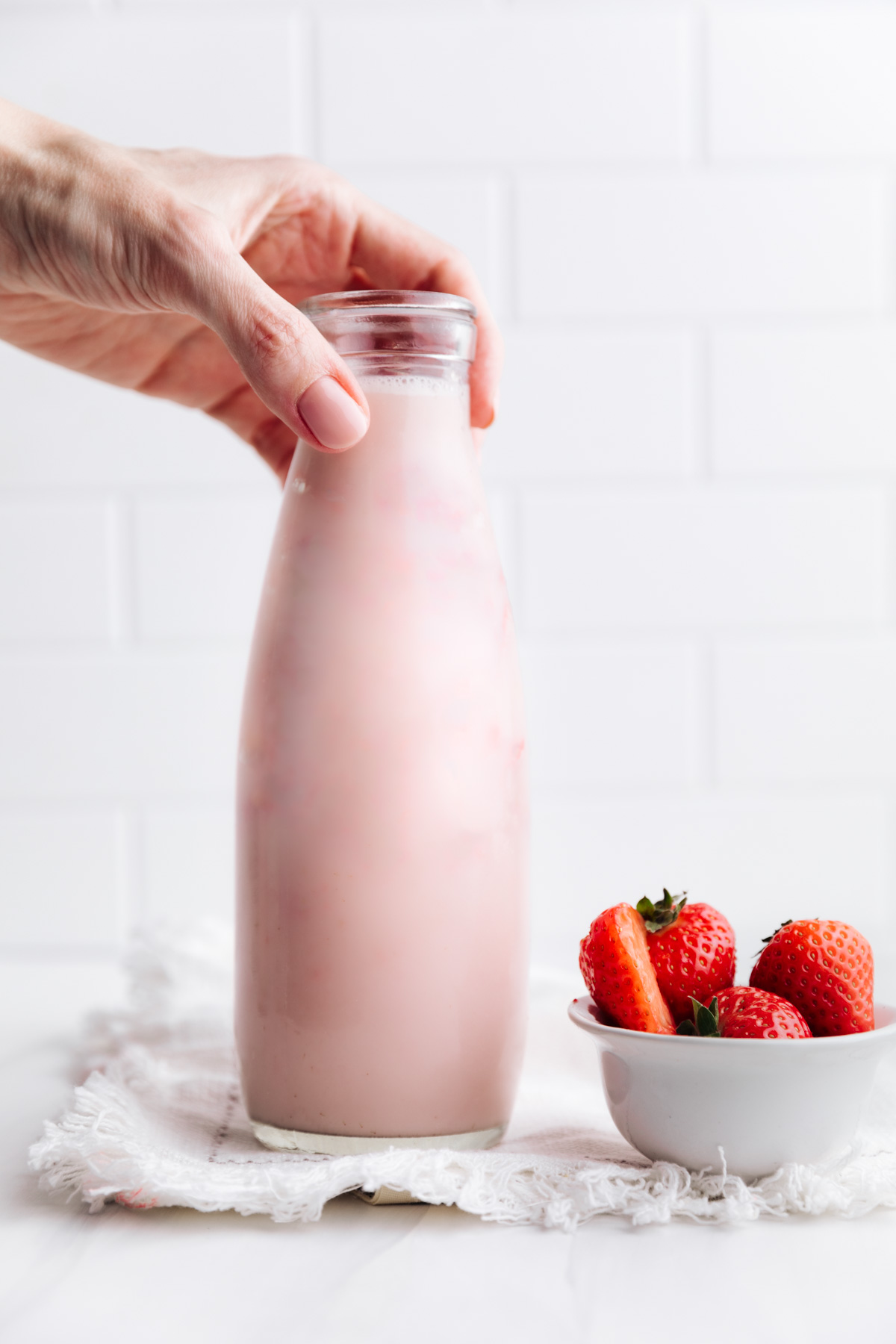 A milk bottle with strawberry milk with a white tiled backdrop and a hand holding the top of the bottle