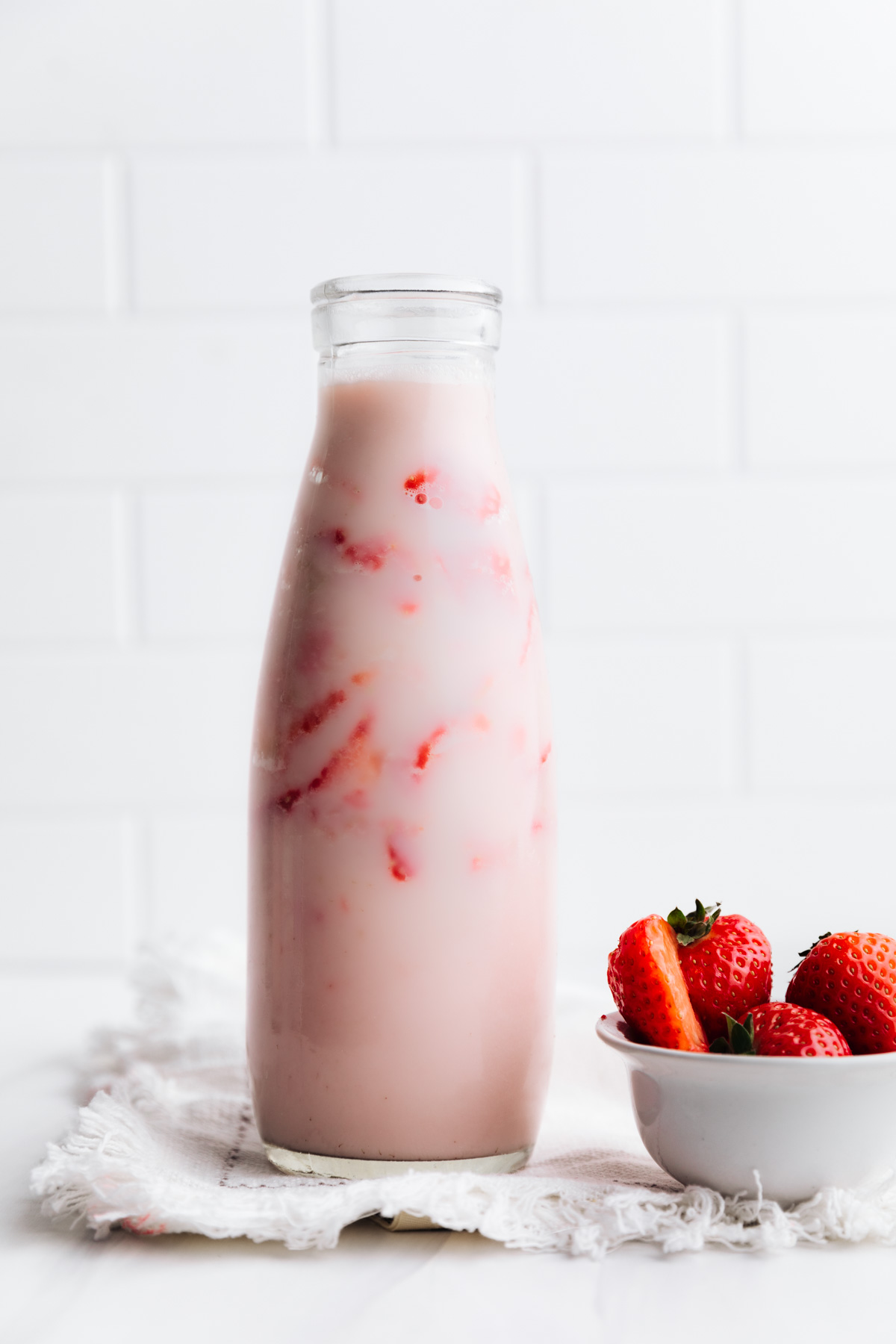 A milk bottle with strawberry milk showing strawberry chunks on a white backdrop next to a small bowl with fresh strawberries