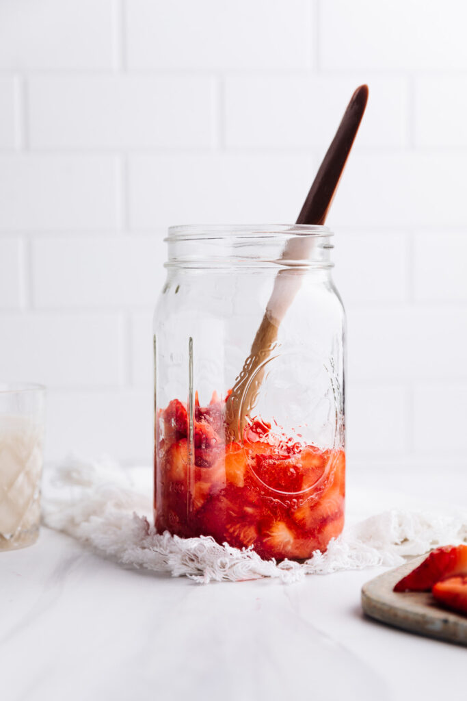 A large glass jar with chopped strawberries, sugar and a wooden spatula in the jar on a white backdrop