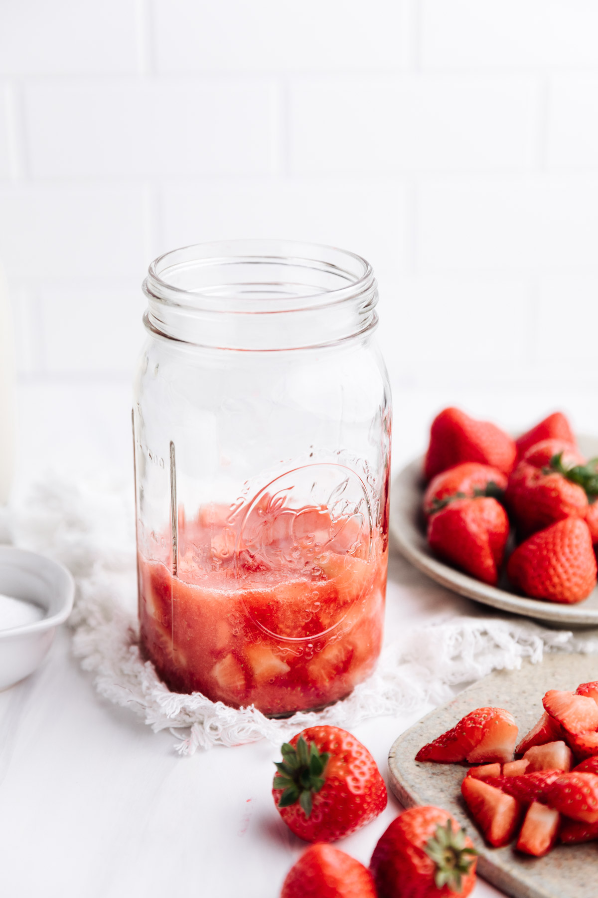 A large glass jar with chopped strawberries and sugar and fresh strawberries in the back in a bowl on a white backdrop