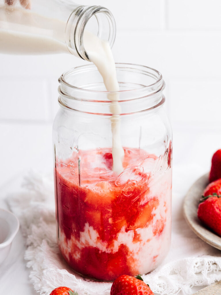 A large glass jar with strawberry and sugar mixture and white milk being poured in with a milk bottle