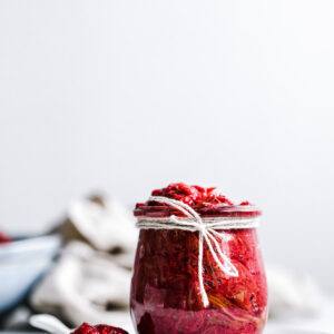 glass jar with red rhubarb jam on a white plate with a teaspoon with rhubarb jam on it with blue bowls in the white background