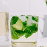 A glass with Hugo Spritz and with a lime wedge, mint leaves and ice cubes in the glass and elderflower decorations around it.