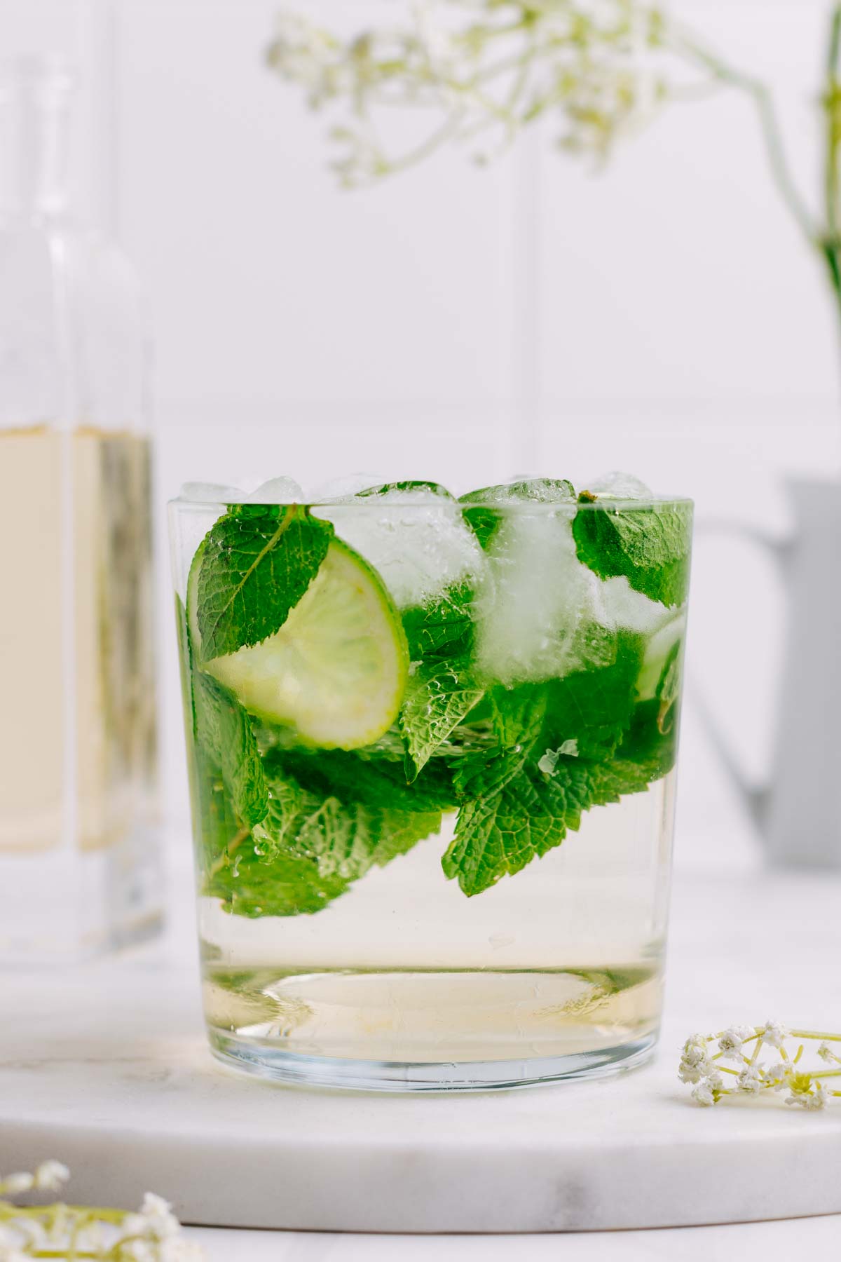 A glass with Hugo Spritz and with a lime wedge, mint leaves and ice cubes in the glass and elderflower decorations around it.