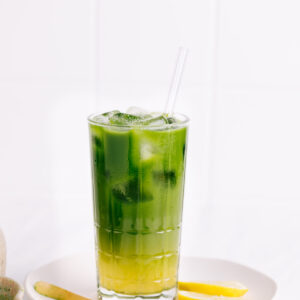 Matcha lemonade in a glass with ice cubes with a separate yellow and green layer with a glass straw on a white plate with lemon slices