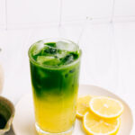A glass with yellow lemonade on the bottom and green matcha tea with ice cubes on the top with a glass straw on a white plate with lemon slices in front of a white kitchen wall