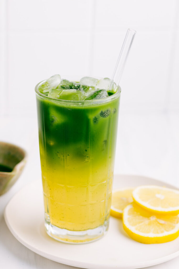 A glass with yellow lemonade on the bottom and green matcha tea with ice cubes on the top with a glass straw on a white plate with lemon slices with a small bowl with green matcha tea in the background