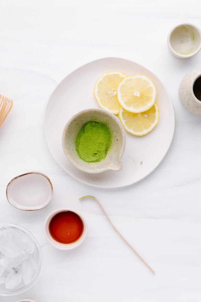 Matcha powder in a small bowl on a white plate with lemon slices on a white backdrop with ingredients for matcha lemonade in small bowls