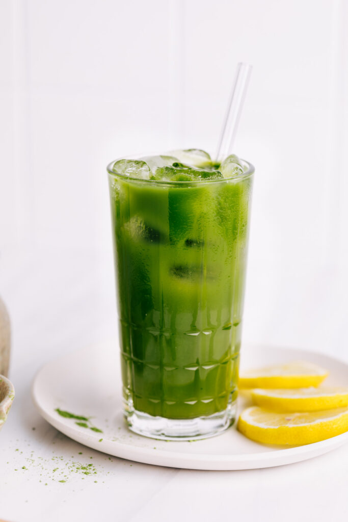 Green colored matcha lemonade in a glass with ice cubes with a glass straw on a white plate with lemon slices and green matcha powder