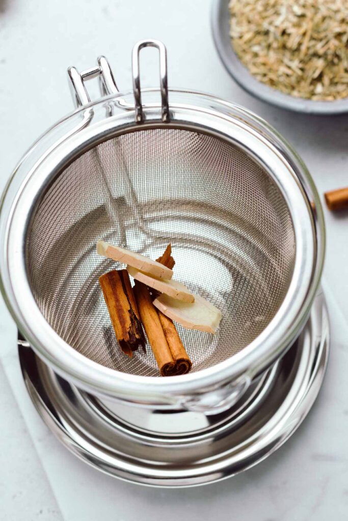 A top view of a tea strainer in a teapot with a broken cinnamon stick and ginger pieces in the strainer.