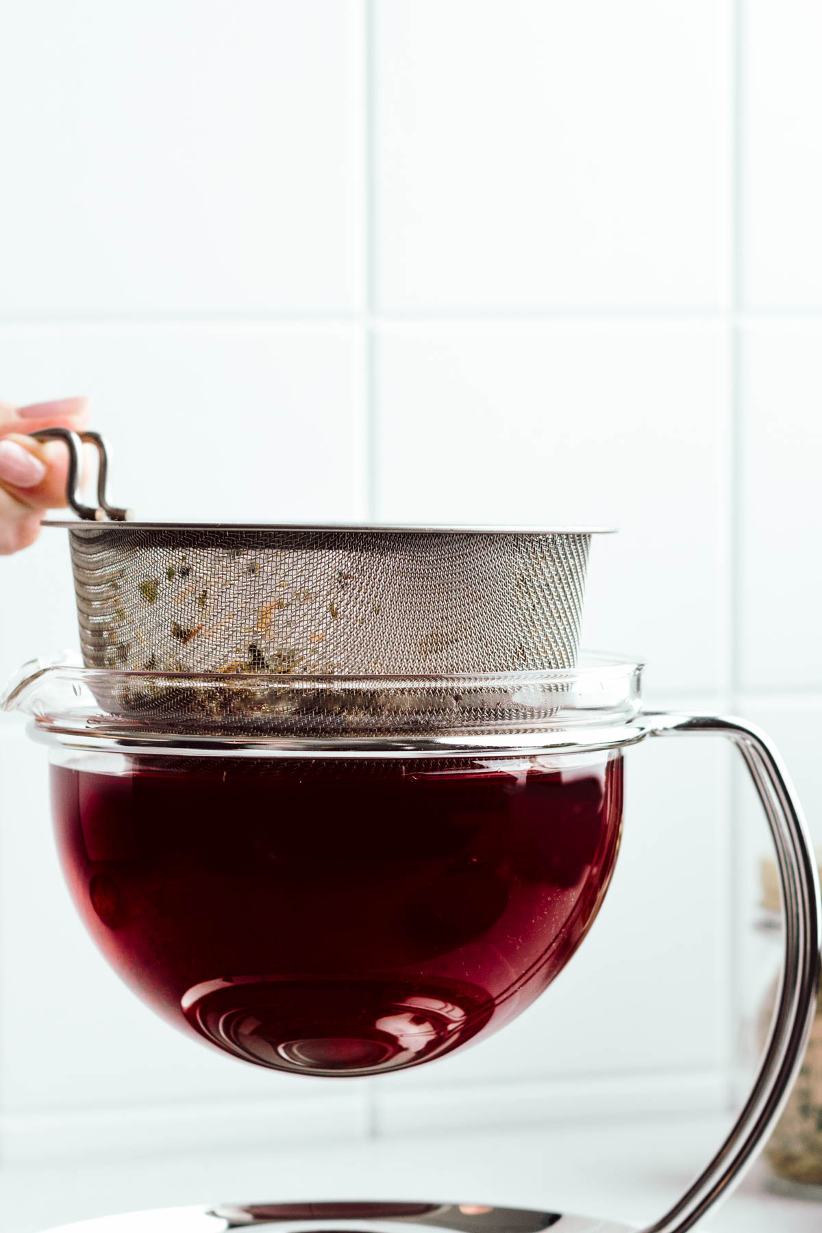 A glass teapot with dark red colored tea in it and a hand lifting the strainer out of the teapot in front of a white tiled backdrop.