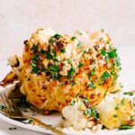 A whole head of golden brown roasted cauliflower covered with nuts and green herbs on a white plate next to a gold fork with slices of cauliflower in front of the head of cauliflower.