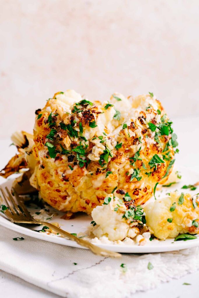 A whole head of golden brown roasted cauliflower covered with nuts and green herbs on a white plate next to a gold fork with slices of cauliflower in front of the head of cauliflower.
